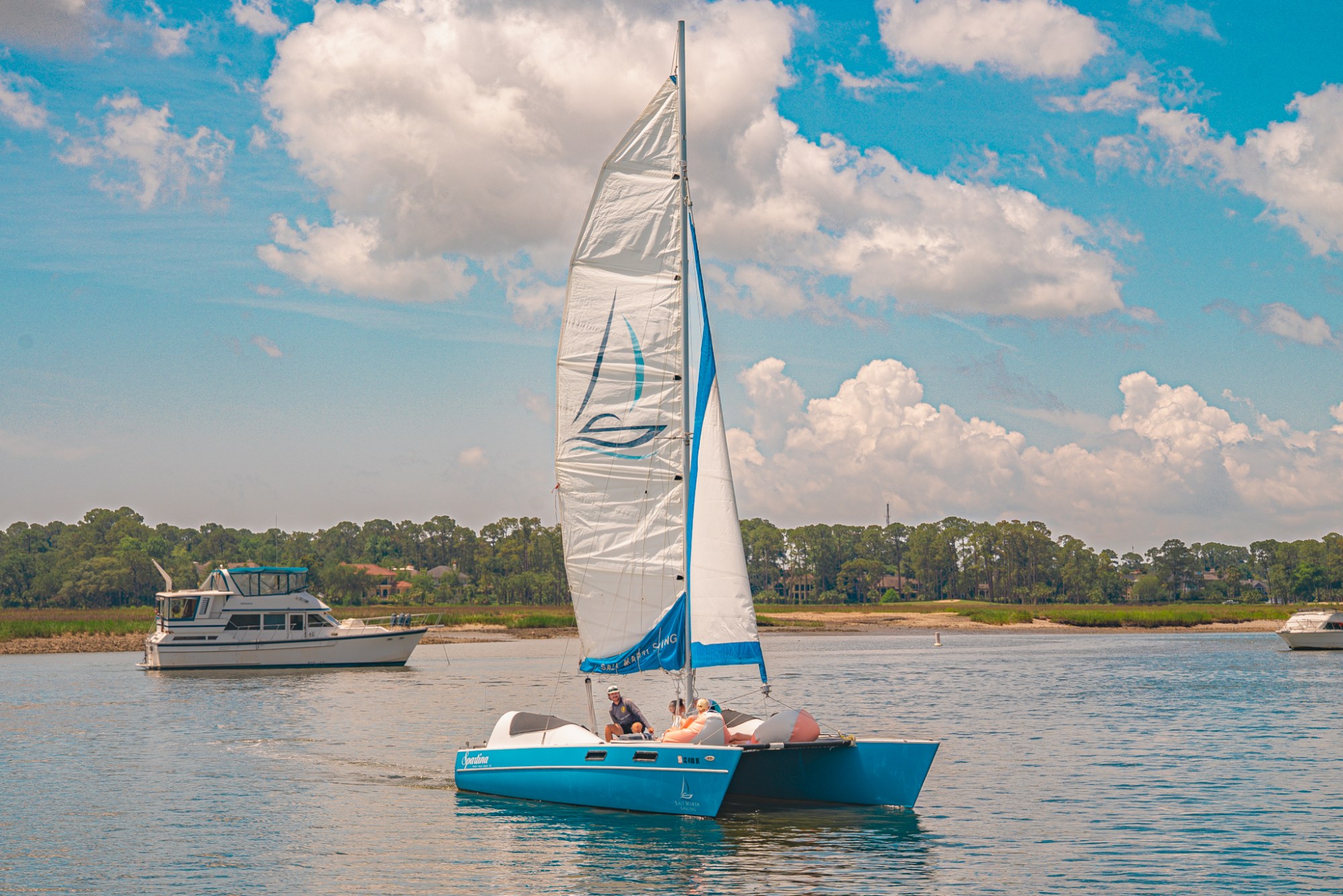 Blue sailboat with white sail on a calm river, trees and another boat in the background.