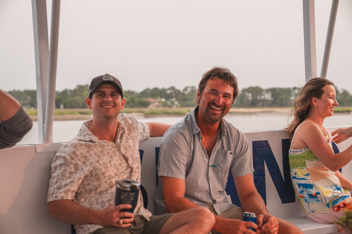 Three people smiling and relaxing on a boat, holding drinks, with water and trees in the background.