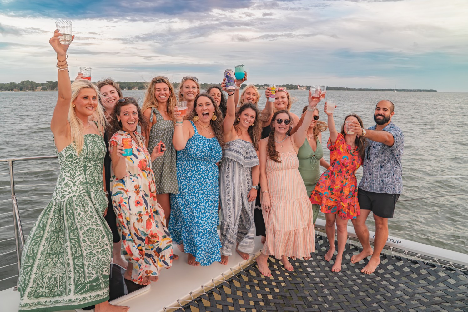 Group of people on a boat, smiling and raising drinks towards the camera with a lake in the background.