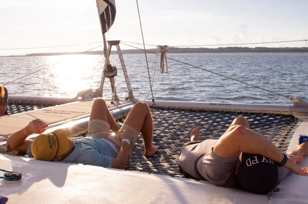 Two people relaxing on a catamaran net with ocean view and sunset.
